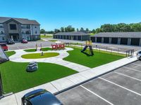 Modern apartment complex with a vibrant playground and ample parking, surrounded by lush greenery under a clear blue sky.