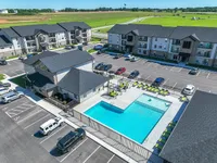 Charming apartment complex with modern buildings, ample parking, and an inviting swimming pool surrounded by sun loungers, set against a backdrop of lush green fields.