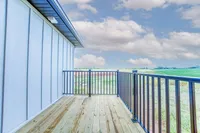 Charming wooden balcony with sleek black railing, offering breathtaking views of lush green fields under a vibrant blue sky.