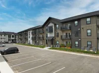 Modern three-story apartment building with balconies, ample parking space, and manicured green surroundings under a clear blue sky.