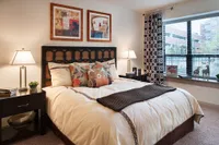 Inviting bedroom featuring a plush bed adorned with floral cushions, elegant bedside lamps, abstract wall art, and a large window with patterned curtains.