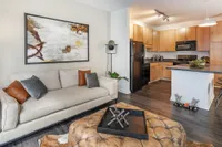 Modern living space featuring a cozy beige sofa, stylish artwork, and an inviting open-concept kitchen with warm wooden cabinets and sleek black appliances.