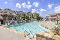 Inviting outdoor pool area with stylish lounge chairs and a shaded pergola, surrounded by beautiful brick buildings and lush greenery under a bright blue sky.