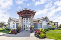 Elegant entrance of a modern apartment complex featuring stone columns, large windows, and vibrant landscaping under a clear blue sky.