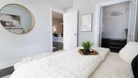 Modern bedroom with a cozy aesthetic, featuring a stylish round mirror, sleek closet space, and inviting decor with a glimpse into a well-lit study area.