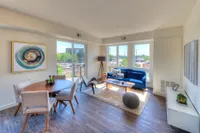 Bright and modern living space featuring a cozy blue sofa, sleek wooden dining table, and large windows with a city view.