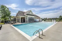 Inviting outdoor swimming pool area with modern patio furniture and pristine clubhouse, surrounded by lush landscaping under a blue sky.
