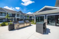 Modern outdoor kitchen area with sleek stainless steel grills and elegant patio seating, set against a backdrop of stylish apartment buildings under a clear blue sky.