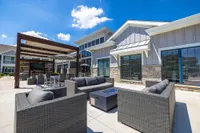 Chic outdoor lounge with modern rattan furniture, shaded pergola, and sleek fire pit, set against a stylish residential building under a clear blue sky.
