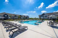 Luxurious apartment complex featuring a sparkling swimming pool surrounded by modern sun loungers, with charming two-story buildings in the background under a clear blue sky.