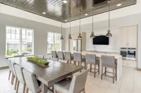 Modern communal dining area with elegant pendant lighting, large wooden table, and chic open-plan kitchen featuring stainless steel appliances and stylish countertops.