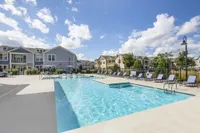 Modern apartment complex featuring a sparkling outdoor pool, surrounded by sun loungers and lush greenery under a bright blue sky.