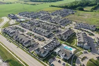 Aerial view of a modern apartment complex featuring multiple buildings with pitched roofs, well-maintained landscaping, ample parking, and a sparkling swimming pool, surrounded by lush green fields.