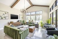 Bright and airy living room with modern decor, featuring green velvet sofas, high ceilings with wooden beams, and large windows overlooking lush greenery.