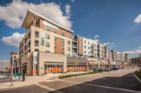 Modern multi-story apartment complex with contemporary design, featuring retail shops on the ground level and balconies overlooking a bustling urban setting.