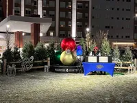 Festive outdoor setup featuring large decorative ornaments, lush greenery, and holiday lights in front of a modern building at Fishers District.