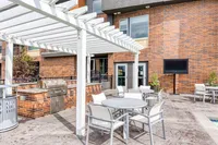 Modern outdoor patio featuring a barbecue grill, elegant seating area with white tables and chairs, and lush greenery against a stylish brick building backdrop.