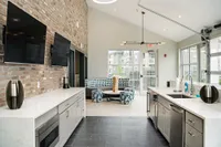 Modern communal area with sleek kitchen, dual TVs mounted on a stylish brick wall, and a cozy seating area with natural light streaming through large windows.