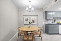 Charming dining area with a round wooden table and stylish chairs, complemented by modern art on a white brick wall, adjacent to a sleek gray kitchen.