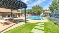 Green grass, brick and cement picnic table under a gazebo in the forground.  Pool with lounge chairs, trees in background.