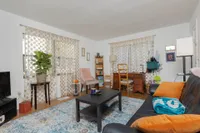 Living room with windows, hardwood floors, white and blue area rug, couch with orange pillows, desk and coffee table.