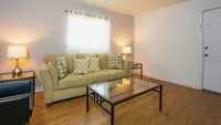 Living area with wood floor, light green fabric covered couch, glass coffee and end tables with lamps.