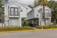 Exterior of the two story Beach House. There is a road in front, a sidewalk leading to the entrance, mailboxes, balconies, and various plants.