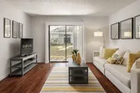 Living room room with wood style flooring, TV, coffee table, and cream colored couch. Sliding glass door overlooks grass exterior.