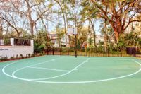 Full size basketball court with fenced in with trees surrounding it.  Tennis court in background..