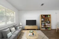 Living room with hardwood floors, contemporary furniture, wall mounted television and wall art. You can see a large window in the background as well as the entrance to the apartment.