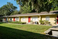 Facade of one level apartment building showing several front doors behind lush green grass and landscaping.