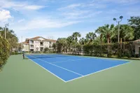 Blue and green tennis court surrounded by fence, bushes and trees. Basketball court in background.