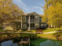 Charming apartment building surrounded by lush greenery and serene pond, featuring a quaint wooden bridge and inviting balconies.