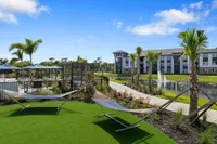 Vibrant apartment complex featuring palm-lined pathways, relaxing hammocks, and peaceful water feature under a bright blue sky.