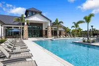 Luxurious apartment complex featuring a sparkling pool surrounded by elegant lounge chairs, lush palm trees, and a modern clubhouse under a clear blue sky.