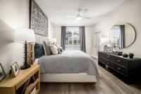 Elegant bedroom featuring a cozy bed with stylish pillows, large window with gray curtains, modern ceiling fan, and sleek black dresser with round mirror.