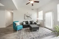 Elegant living room with a modern gray sofa, teal chair, and stylish decor, featuring a fan and ample natural light through large windows.