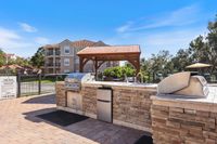 Charming outdoor grilling area with a modern BBQ setup under a stylish wooden gazebo, overlooking a well-maintained residential complex with lush greenery and clear blue skies.