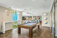 Luxurious apartment lounge with contemporary decor, featuring a pool table, modern lighting, and spacious seating area with a view of palm trees through large windows.