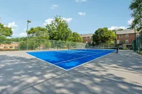 Vibrant blue tennis court surrounded by lush greenery and charming brick apartments, offering a perfect blend of recreation and community living.