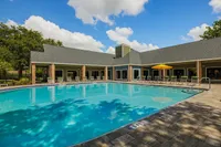 Inviting outdoor pool area with modern deck chairs and vibrant yellow umbrellas, surrounded by lush greenery and a beautiful clubhouse under a bright blue sky.