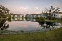 Charming lakeside apartments with serene reflections, surrounded by lush greenery and a picturesque sunset sky.