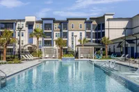Luxurious apartment complex with a sparkling pool, surrounded by loungers and palm trees, under a clear blue sky.
