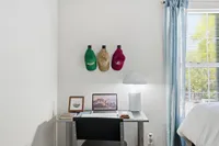 Modern workspace in bright apartment bedroom featuring a sleek desk with a laptop, contemporary lamp, and decor accents beneath colorful hats. Sunlit window with soft blue curtains adds a cozy touch.