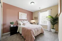 Inviting bedroom with cozy decor featuring soft pink and beige tones, large window with sheer curtains, and lush potted plant adding a touch of nature.