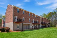 Charming brick apartment building with individual balconies, surrounded by lush green lawns and scenic trees, perfect for serene living.