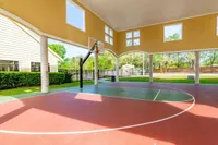 Inviting covered basketball court with vibrant red and green flooring, surrounded by lush greenery and ample natural light.