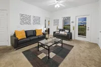 Inviting living room with modern decor, featuring sleek black leather sofas, vibrant orange cushions, and artistic wall hangings. Bright natural light streams through large windows onto a stylish glass coffee table and patterned carpet.