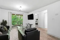 Elegant living room with modern black leather sofas, a flat screen TV, and a view of lush green trees through large glass doors leading to a balcony.