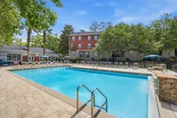 Inviting outdoor pool area with sun loungers, surrounded by lush greenery and charming brick buildings under a clear blue sky.